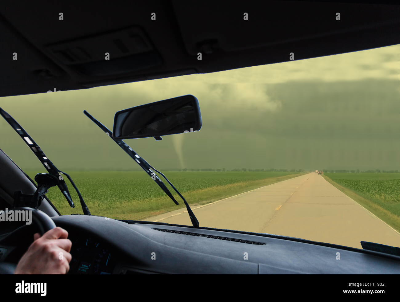 collage from two stock images. Perspective from passenger side of car, where driver's hand can be seen on steering wheel. Windshield view is of a lone highway cutting through a field, with a tornado in the distance